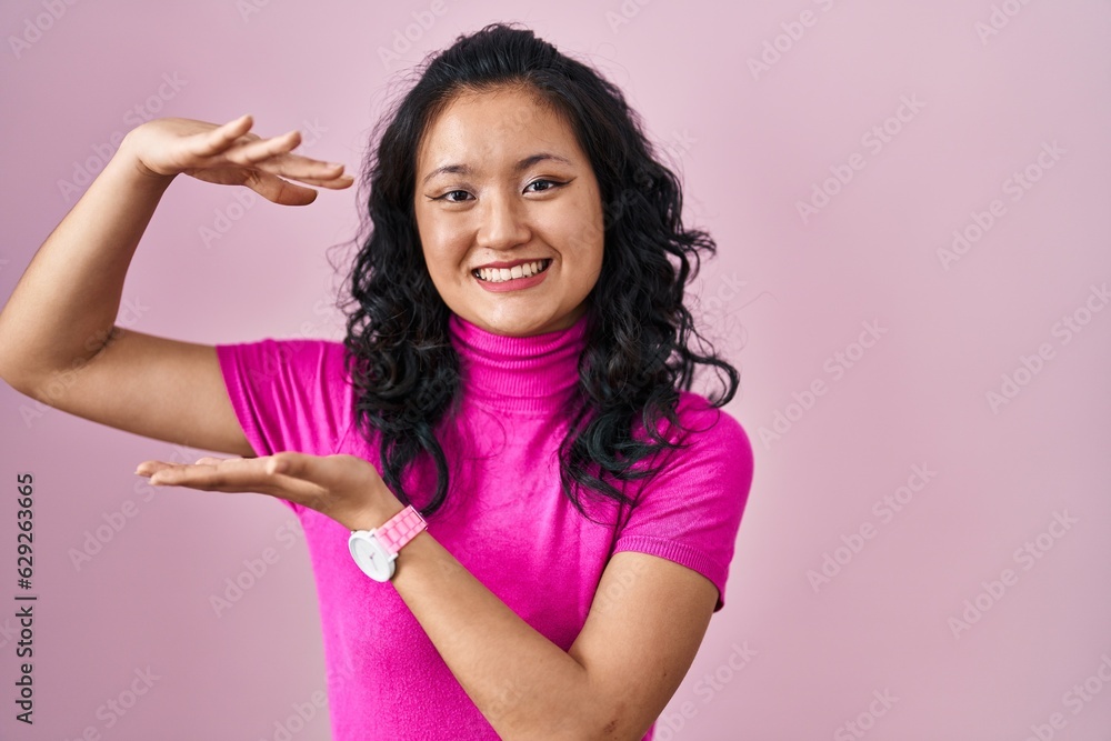 Sticker young asian woman standing over pink background gesturing with hands showing big and large size sign