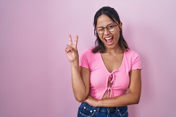Hispanic young woman standing over pink background wearing glasses smiling with happy face winking at the camera doing victory sign. number two.