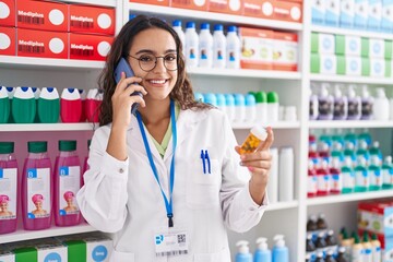 Young beautiful hispanic woman pharmacist holding pills bottle talking on smartphone at pharmacy