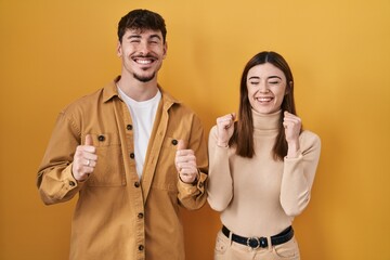 Young hispanic couple standing over yellow background excited for success with arms raised and eyes closed celebrating victory smiling. winner concept.