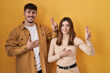 Young hispanic couple standing over yellow background smiling swearing with hand on chest and...