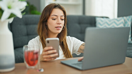Young beautiful hispanic woman using laptop and smartphone sitting on floor at home