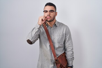 Young hispanic man wearing suitcase mouth and lips shut as zip with fingers. secret and silent,...
