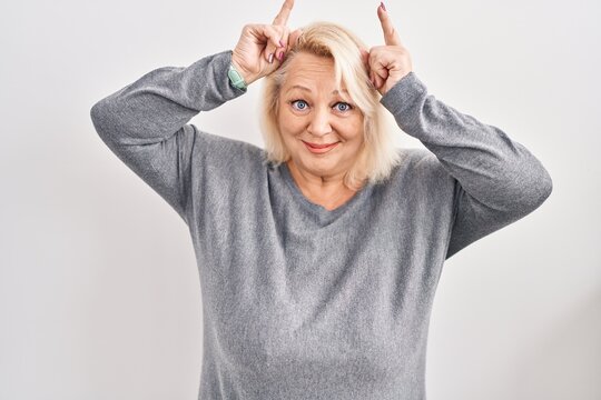 Middle age caucasian woman standing over white background doing funny gesture with finger over head as bull horns