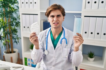 Young doctor man holding two safety mask clueless and confused expression. doubt concept.