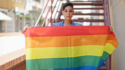 Young beautiful hispanic woman standing with serious expression holding rainbow flag at street