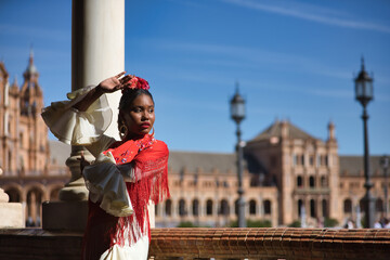 Young black and South American woman in a beige gypsy flamenco suit and red shawl, dancing in a beautiful square in the city of Seville in Spain. Concept dance, folklore, flamenco, art.