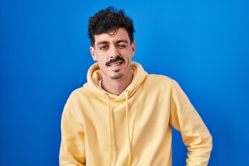 Hispanic man standing over blue background winking looking at the camera with sexy expression, cheerful and happy face.