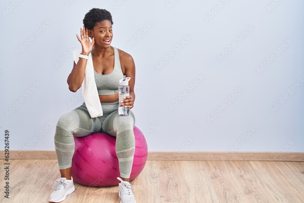 Wall mural African american woman wearing sportswear sitting on pilates ball smiling with hand over ear listening an hearing to rumor or gossip. deafness concept.