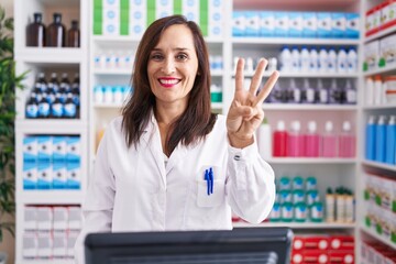 Middle age brunette woman working at pharmacy drugstore showing and pointing up with fingers number three while smiling confident and happy.