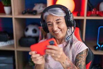 Middle age grey-haired woman streamer playing video game using smartphone at gaming room