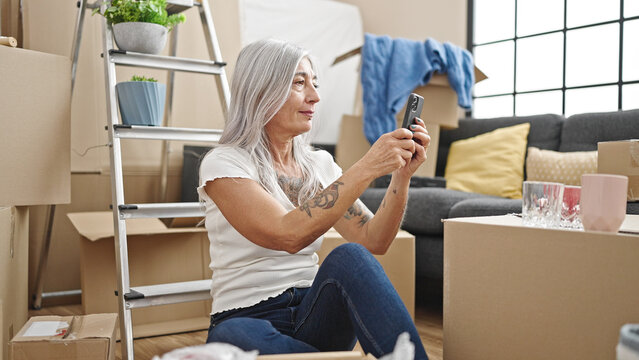 Middle age grey-haired woman recording video by smartphone sitting on floor at new home