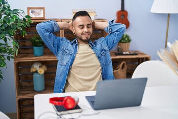 Young hispanic man sitting on table relaxed with hands on head at home