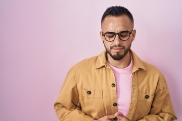 Obraz premium Young hispanic man standing over pink background with hand on stomach because indigestion, painful illness feeling unwell. ache concept.