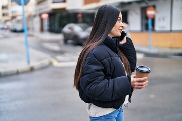 Young beautiful hispanic woman talking on smartphone drinking coffee at street