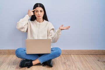 Young woman using laptop sitting on the floor at home confused and annoyed with open palm showing copy space and pointing finger to forehead. think about it.