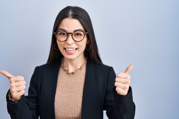 Young brunette woman standing over blue background success sign doing positive gesture with hand, thumbs up smiling and happy. cheerful expression and winner gesture.