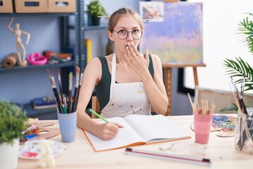 Young caucasian woman sitting at art studio painting on notebook covering mouth with hand, shocked...