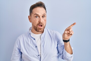 Middle age caucasian man standing over blue background surprised pointing with finger to the side, open mouth amazed expression.