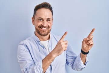 Middle age caucasian man standing over blue background smiling and looking at the camera pointing with two hands and fingers to the side.