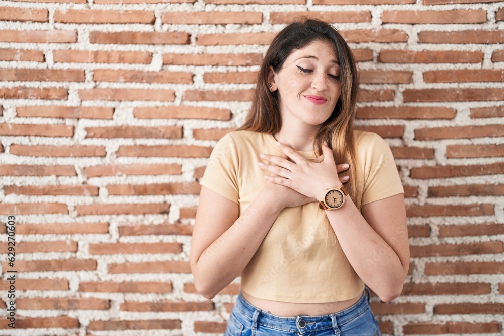 Sticker Young brunette woman standing over bricks wall smiling with hands on chest with closed eyes and grateful gesture on face. health concept.