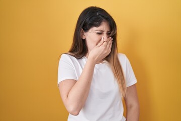 Young brunette woman standing over yellow background smelling something stinky and disgusting, intolerable smell, holding breath with fingers on nose. bad smell