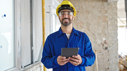 Young hispanic man worker wearing hardhat using touchpad at construction site