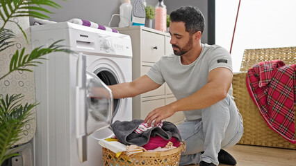 Young hispanic man washing clothes at laundry room - obrazy, fototapety, plakaty