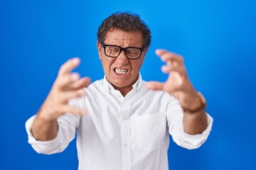 Naklejka premium Middle age hispanic man standing over blue background shouting frustrated with rage, hands trying to strangle, yelling mad