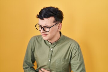 Non binary person standing over yellow background with hand on stomach because nausea, painful disease feeling unwell. ache concept.