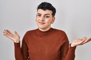 Non binary person wearing make up standing over isolated background clueless and confused expression with arms and hands raised. doubt concept.