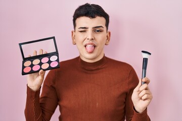 Non binary person holding makeup brush and blush sticking tongue out happy with funny expression.