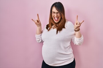Pregnant woman standing over pink background shouting with crazy expression doing rock symbol with hands up. music star. heavy music concept.
