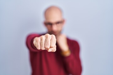 Young bald man with beard standing over white background wearing glasses punching fist to fight, aggressive and angry attack, threat and violence