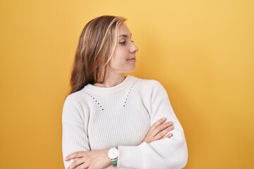 Young caucasian woman wearing white sweater over yellow background looking to the side with arms crossed convinced and confident