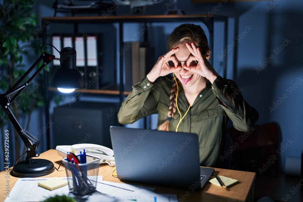 Sticker Young blonde woman working at the office at night doing ok gesture like binoculars sticking tongue out, eyes looking through fingers. crazy expression.