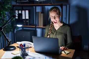 Young blonde woman working at the office at night winking looking at the camera with sexy...