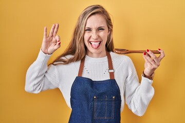 Young blonde woman wearing apron tasting food holding wooden spoon sticking tongue out happy with...