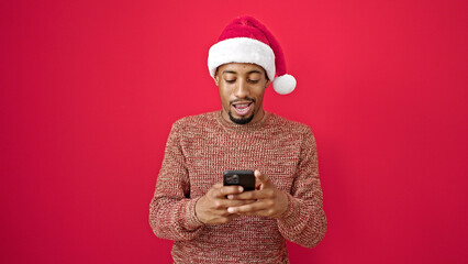 African american man wearing christmas hat using smartphone smiling over isolated red background