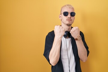 Young caucasian man wearing sunglasses standing over yellow background ready to fight with fist defense gesture, angry and upset face, afraid of problem