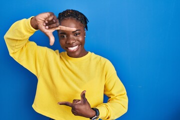 Beautiful black woman standing over blue background smiling making frame with hands and fingers with happy face. creativity and photography concept.