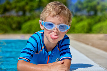 A funny Caucasian boy with swimming goggles teases on camera near the pool. Positive baby lying on...