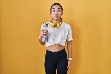 Young south asian woman wearing sportswear drinking water puffing cheeks with funny face. mouth...
