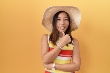 Middle age chinese woman wearing summer hat over yellow background looking confident at the camera smiling with crossed arms and hand raised on chin. thinking positive.