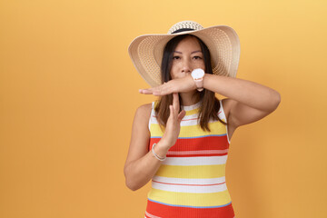Middle age chinese woman wearing summer hat over yellow background doing time out gesture with...