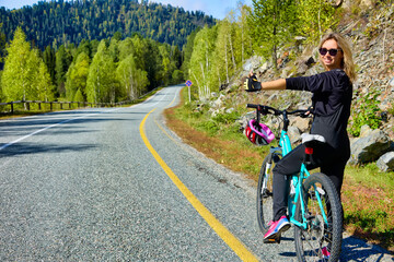 Pretty young girl in sunglasses and black tracksuit stands on side of road on a bicycle, voting with her hand. Girl athlete stopped on the side of the road with a bicycle, stops passing traffic