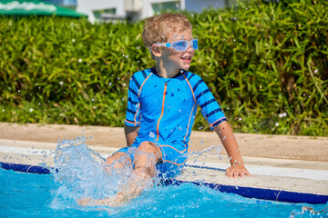 Against backdrop of green bushes, cheerful Caucasian boy in swimming glasses sits by pool...