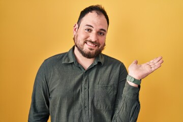 Plus size hispanic man with beard standing over yellow background smiling cheerful presenting and pointing with palm of hand looking at the camera.