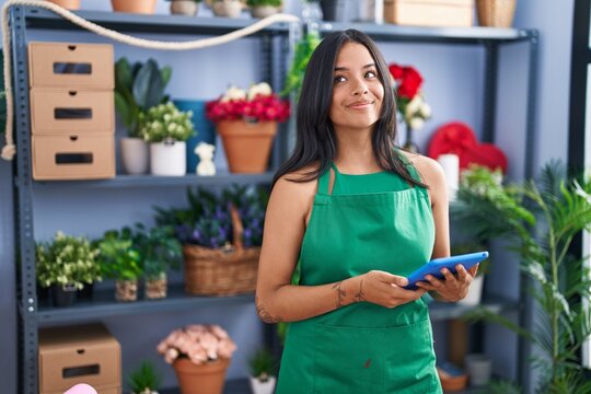 Brunette woman working at florist shop holding tablet smiling looking to the side and staring away thinking.
