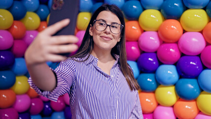 Fototapeta na wymiar Young beautiful hispanic woman visiting colorful futuristic exhibition space taking selfie